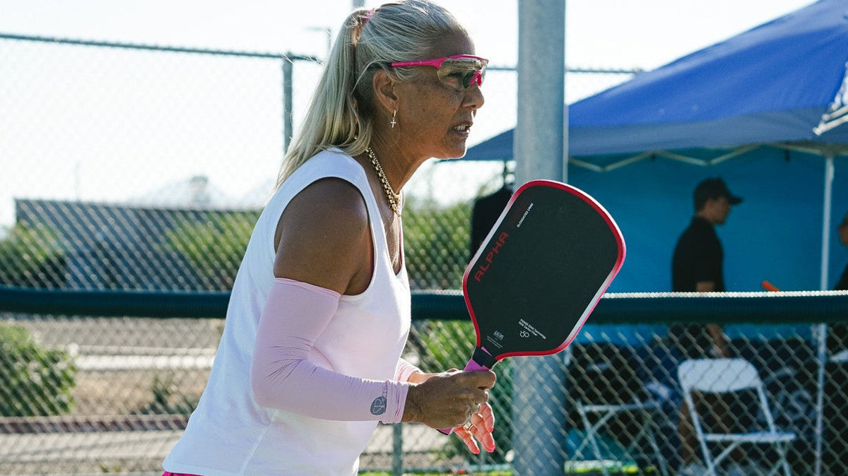 Anna Shirley wearing pink PBPRO Infinity eyewear and holding a paddle beside a court net