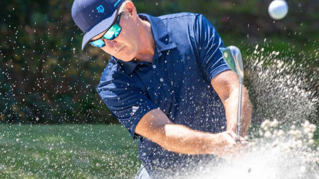 Dan Jansen wearing sunglasses while hitting a golf shot from a water hazard