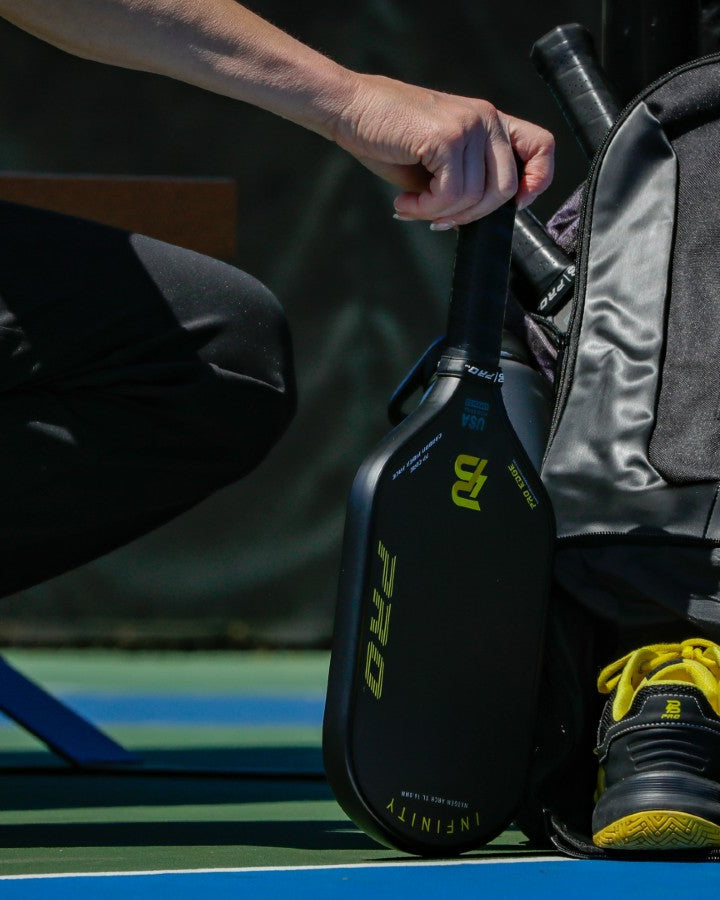 Player placing a black and yellow PBPRO Infinity paddle beside a Tour backpack and matching shoes