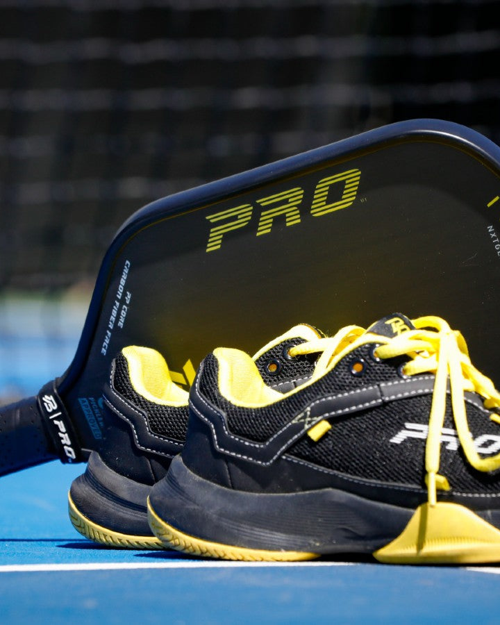 Black and yellow PBPRO Infinity pickleball paddle beside matching shoes on a court