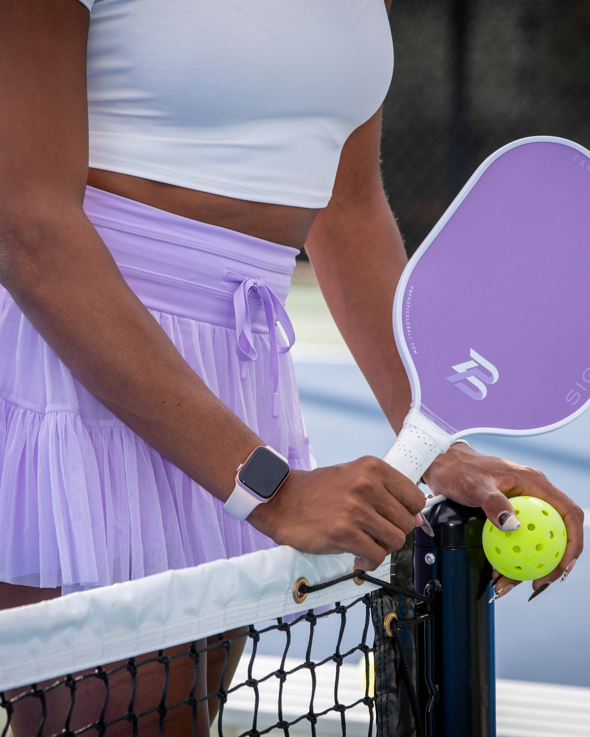 Player in a lavender skirt holding a lavender PB Signature paddle and a pickleball beside a net