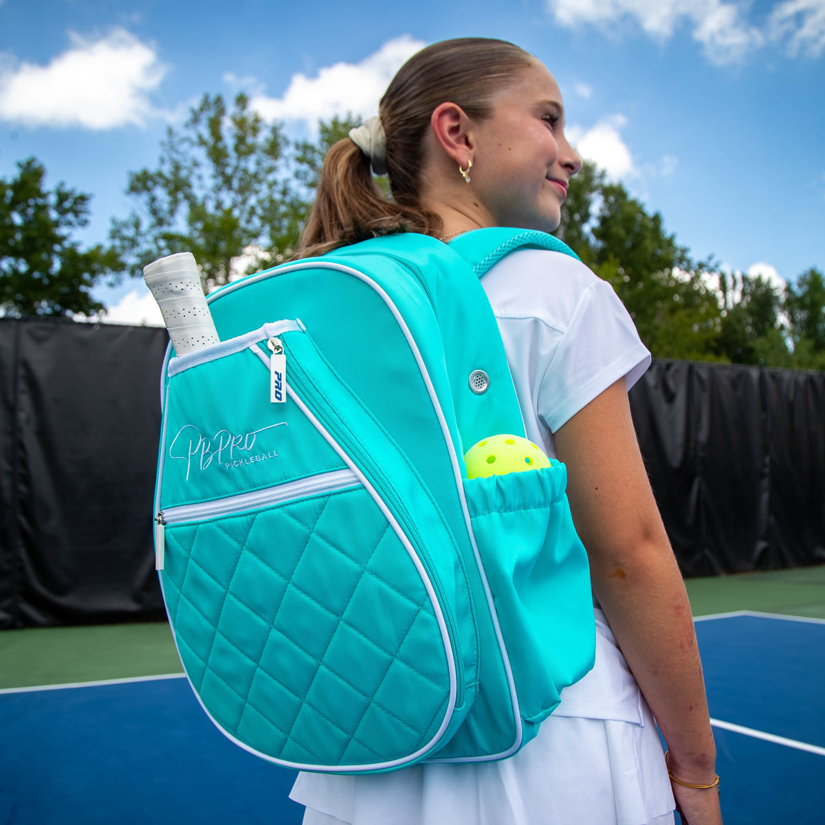 Mint PBPRO Signature pickleball backpack worn on court, with a paddle in the front compartment and ball in the side pocket