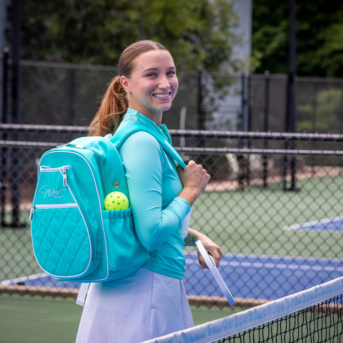 Player carrying a mint PBPRO Signature pickleball backpack beside an outdoor court net
